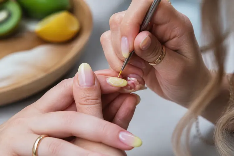 Une jeune femme réalise un nail art estival simple avec des motifs fruités sur un ongle, dans un salon de manucure cosy.