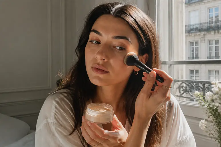 Une jeune femme applique de la poudre Laura Mercier dans un appartement parisien baigné de lumière naturelle.