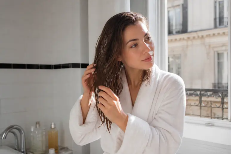 Une femme applique une crème capillaire sur les longueurs et pointes de ses cheveux humides dans une salle de bain lumineuse, après sa douche matinale.