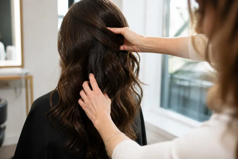 Les mains expertes d'une coiffeuse française touchent délicatement une longue chevelure brune et ondulée dans un salon moderne.