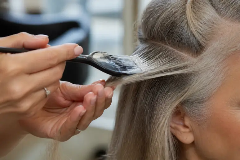 Mains d'une coiffeuse appliquant délicatement un produit éclaircissant sur une mèche de cheveux gris.